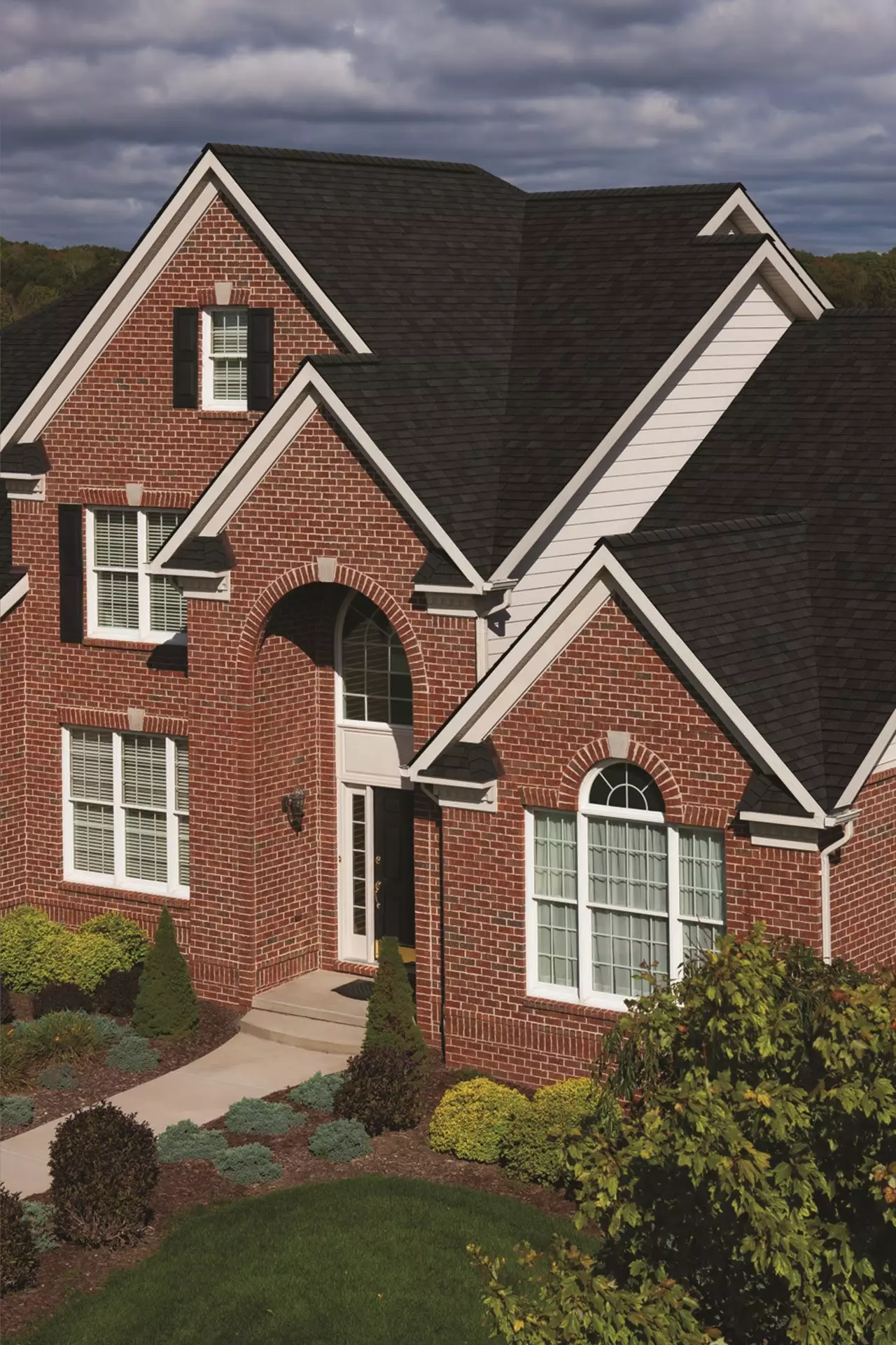 Elegant brick home with dark shingles, white trim, arched windows, and manicured landscaping under cloudy sky.