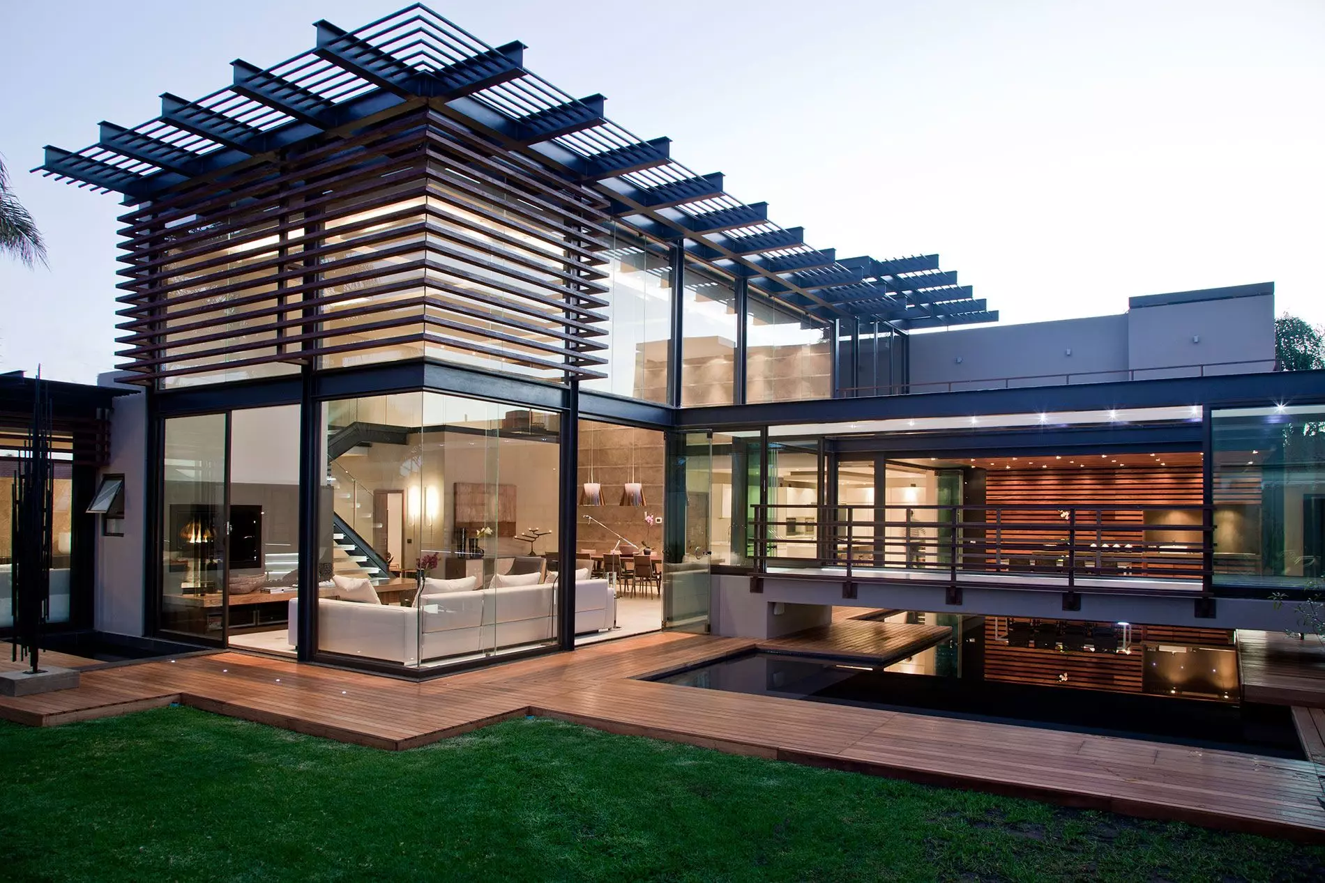 Modern glass house with wooden slat screens, geometric pergola, outdoor deck, and illuminated interior at dusk.